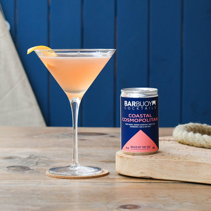 A cocktail in a martini glass with a orange twist sits on a wooden table next to a can of Bar Buoy Cocktails Coastal Cosmopolitan.