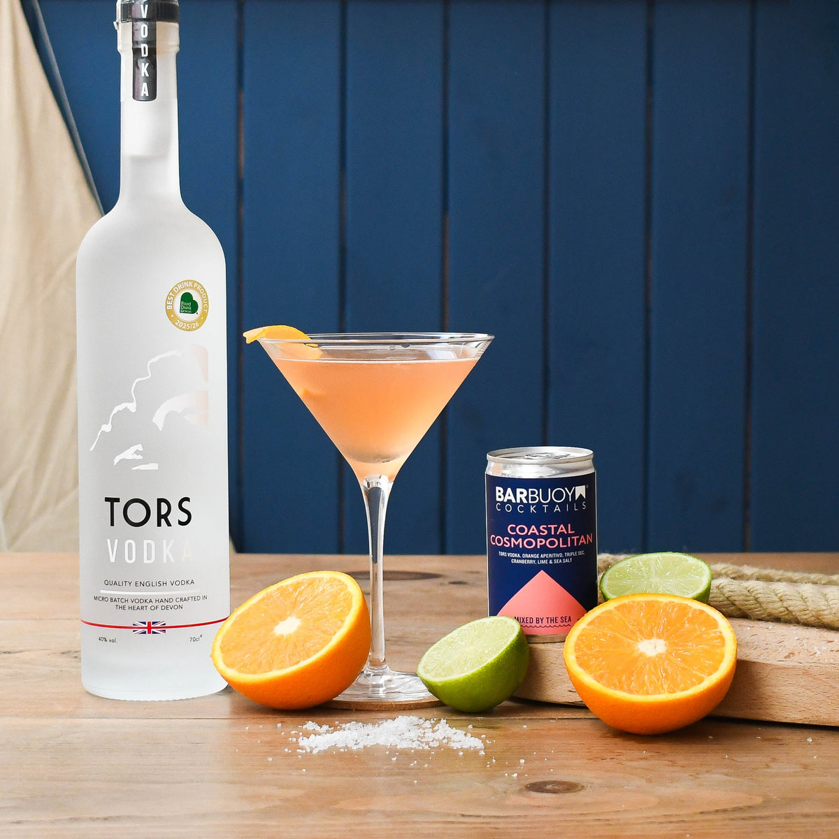 A bottle of Tors Vodka stands on a wooden table beside a filled martini glass with an orange garnish, a can of Bar Buoy Coastal Cosmopolitan, and sliced oranges and limes against a dark blue paneled background.