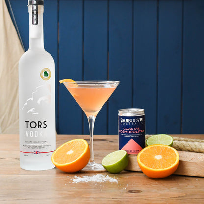 A bottle of Tors Vodka stands on a wooden table beside a filled martini glass with an orange garnish, a can of Bar Buoy Coastal Cosmopolitan, and sliced oranges and limes against a dark blue paneled background.