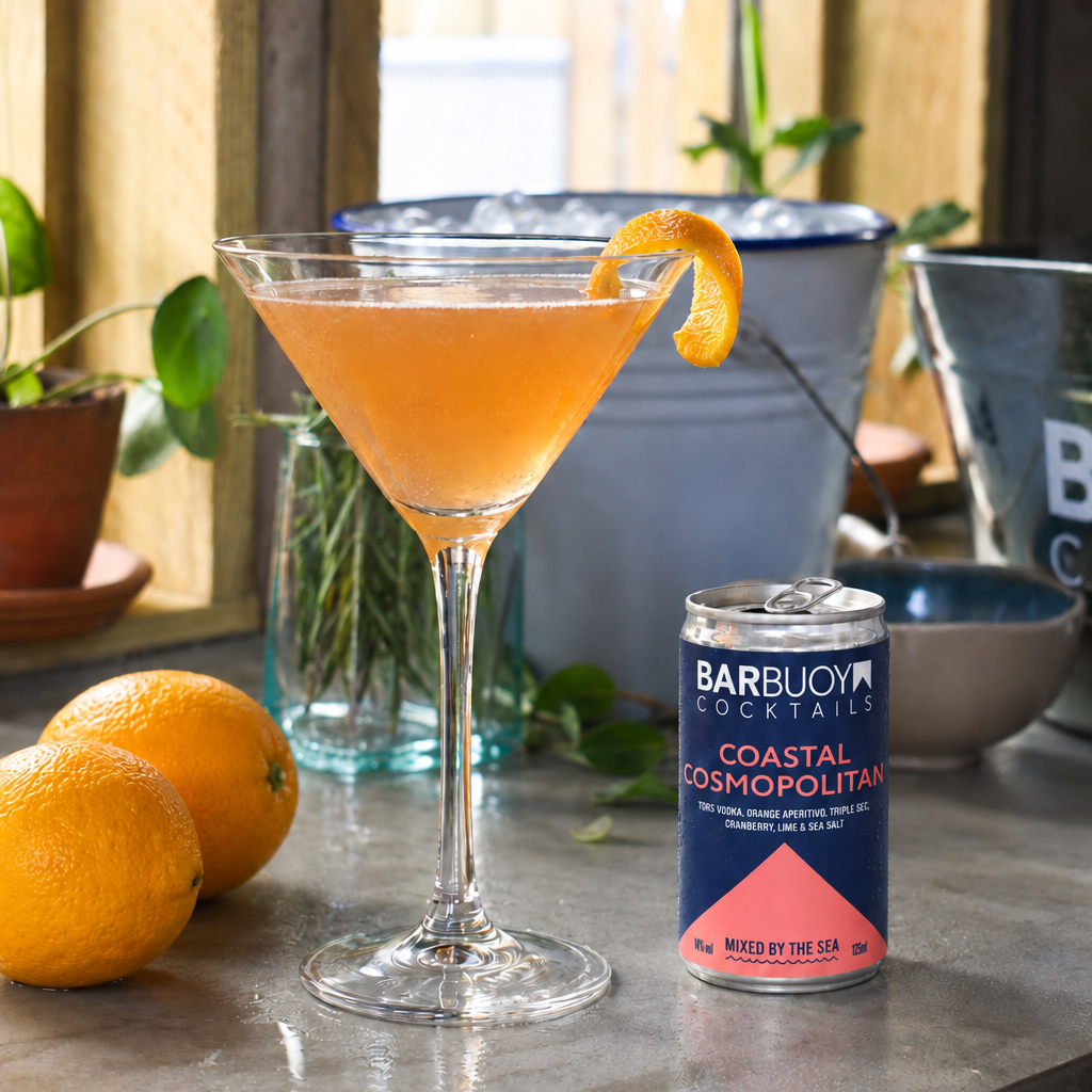 A Bar Buoy Cocktails can labeled Coastal Cosmopolitan stands beside a martini glass filled with orange / pink cocktail liquid and garnished with an orange peel, set on a stone countertop with oranges and bar accessories in a kitchen-like environment.