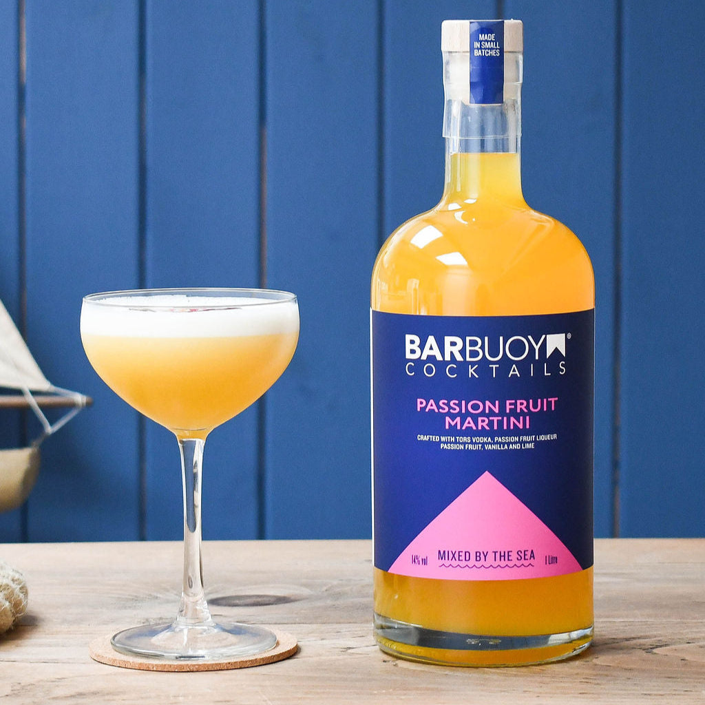 Barbuoy Cocktails Passion Fruit Martini bottle with a glass of the cocktail on a wooden surface against a blue background